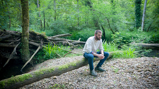 Young man sitting on a fallen log in the middle of the forest by a river