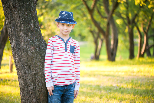 A young boy in a blue hat stands in the garden near the trees and looks straight. Close-up portrait.