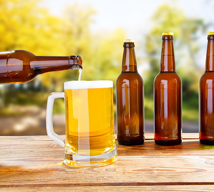bottle pours beer in cup on wooden table on blurred park background