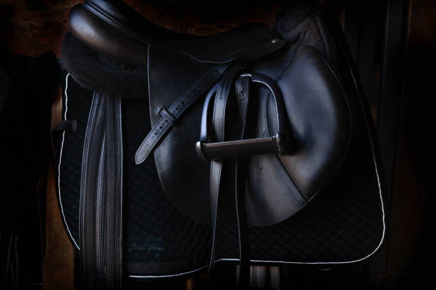 silla de caballo de cuero negro brillante en el establo oscuro, con estribo, listo para montar - saddle fotografías e imágenes de stock