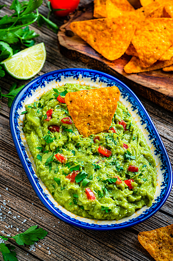 Mexican food: guacamole dipping sauce in a bowl with tortilla chips shot on rustic wooden table. Ingredients for preparing guacamole are all around the bowl and includes ripe avocados, tomatoes, lime, cilantro and salt. Predominant color is green. High resolution 42Mp studio digital capture taken with Sony A7rII and Sony FE 90mm f2.8 macro G OSS lens