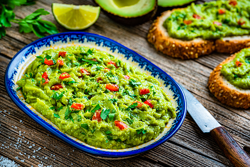 Healthy food: guacamole dipping sauce in a bowl and sliced bread with guacamole shot on rustic wooden table. Ingredients for preparing guacamole are all around the bowl and includes ripe avocados, lime, cilantro and salt. Predominant color is green. High resolution 42Mp studio digital capture taken with Sony A7rII and Sony FE 90mm f2.8 macro G OSS lens