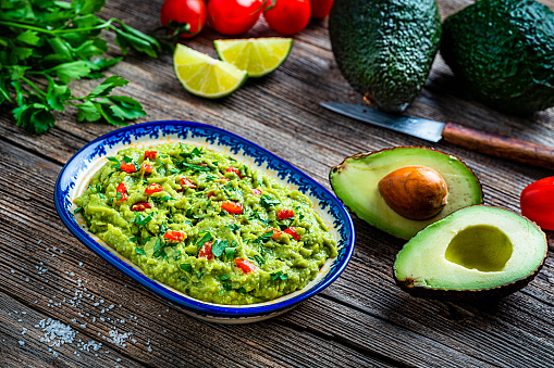 Healthy food: guacamole dipping sauce in a bowl shot on rustic wooden table. Ingredients for preparing guacamole are all around the bowl and includes ripe avocados, tomatoes, lime, cilantro and salt. Predominant color is green. High resolution 42Mp studio digital capture taken with Sony A7rII and Sony FE 90mm f2.8 macro G OSS lens