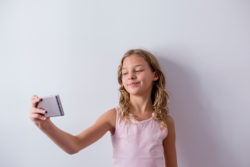 portrait of a young beautiful kid using a mobile phone and taking a selfie. white background. Kids indoors. Lifestyle