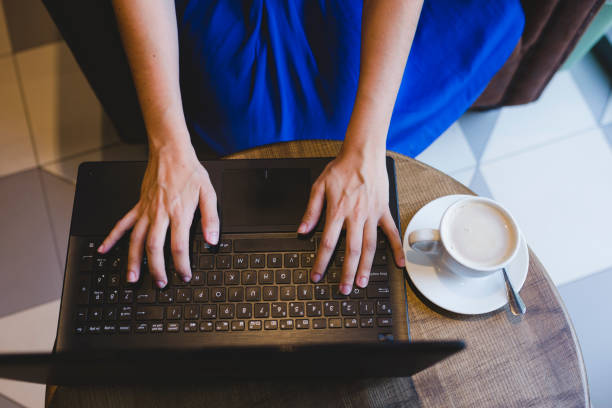 ラップトップ、コーヒーに書いている若い美しい女性のトップビュー。カジュアルな青いドレス。屋内の現代生活 - computer key internet cafe coffee internet ストックフォトと画像