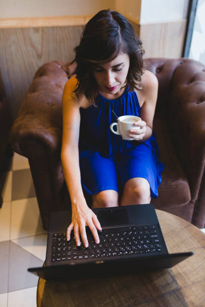 カフェでコーヒーを楽しむ若い美しい女性。ラップトップその他。カジュアルな青いドレス。屋内の現代生活 - computer key internet cafe coffee internet ストックフォトと画像