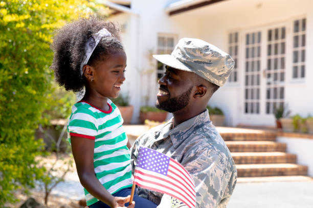 African American male soldier holding his daughter with a US flag in his arms African American male solider wearing uniform holding his daughter with a USA flag in his arms, standing by the house on a sunny day, smiling and interacting. black military man stock pictures, royalty-free photos & images