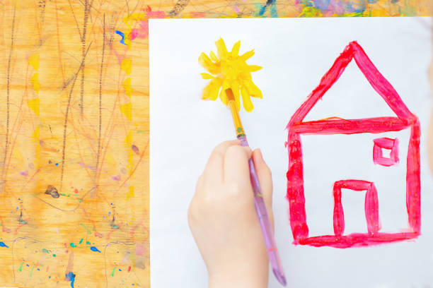 Hand of child drawing sun with red house on white paper on easel. Children creativity.