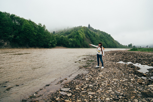 Tourist girl in jeans and white sweatshirt with orange backpack on the bank of the mountain river surrounded by forest, enjoying silence and harmony of nature early in the morning.
