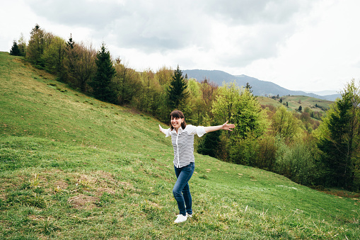 Happy smiling tourist girl up in mountains surrounded by forest, raising arms and enjoying silence and harmony of nature.