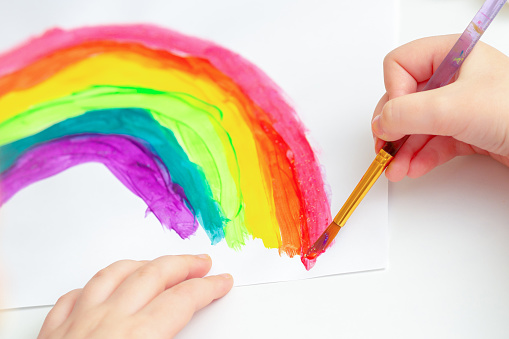 Close up of hands of child drawing rainbow by watercolors on white paper.