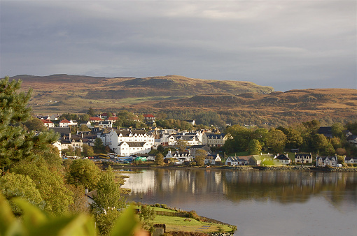 A morning view of Portree Village on the Isle of Skye in western Scotland with rolling hills in the background and the village reflecting in the water