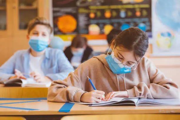 High school students at school, wearing N95 Face masks. Teenage girl wearing eyeglasses sitting at the school desk and writing int he notebook.