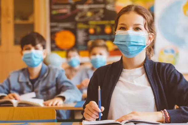 High school students at school, wearing N95 Face masks. Teenage girl sitting at the school desk and listening to the teacher.