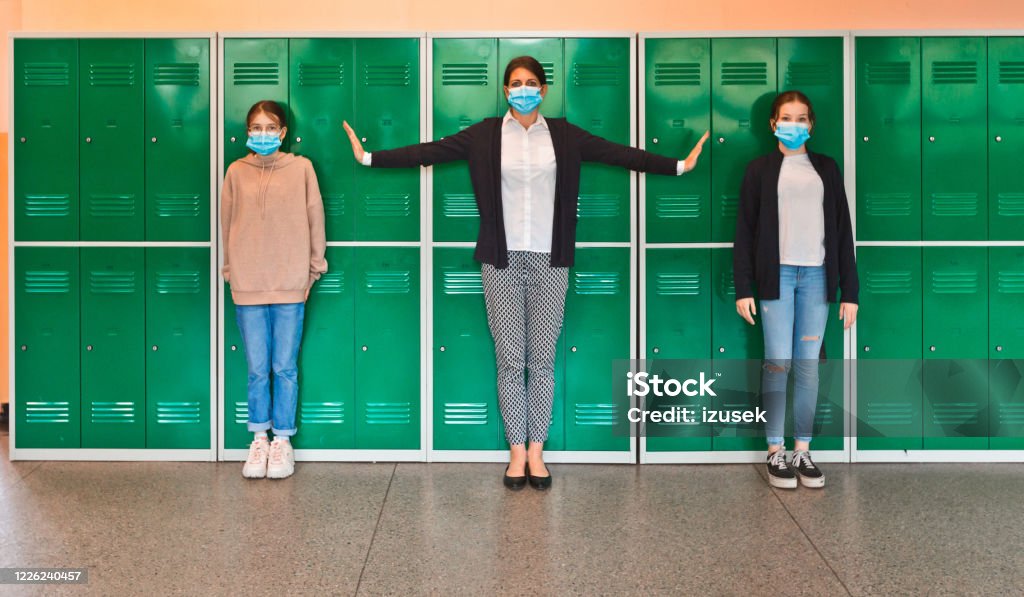 High school students and teacher wearing N95 Face masks at school Teenage girls and female teacher wearing N95 face masks standing in front of school lockers. Woman outstretching arms in the distance. High school students at school. Back to School Stock Photo