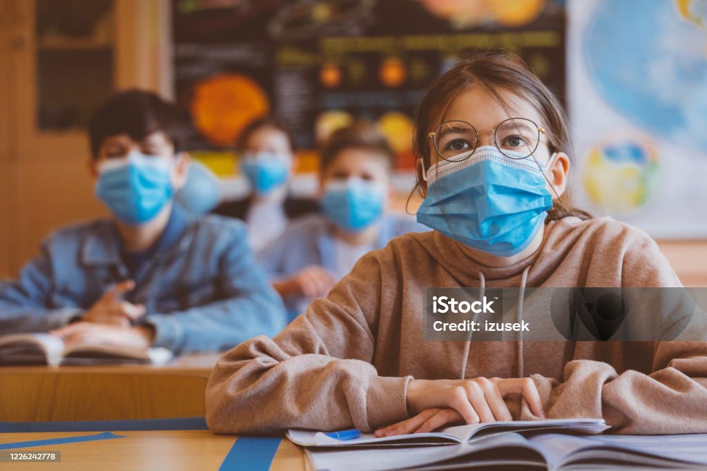 Teenage girl wearing N95 face mask at school High school students at school, wearing N95 Face masks. Teenage girl wearing eyeglasses sitting at the school desk and listening to the teacher. Protective Face Mask Stock Photo