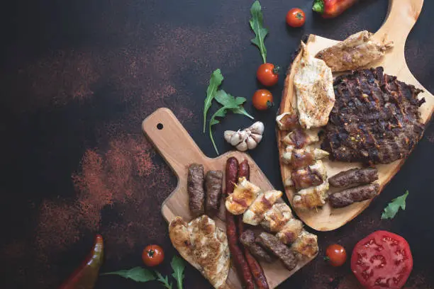 Various grilled meats served on wooden cutting boards with fresh vegetables and bread on a dark rustic background. Traditional Serbian and Balkan bbq meat called rostilj. Flat lay. Copy space