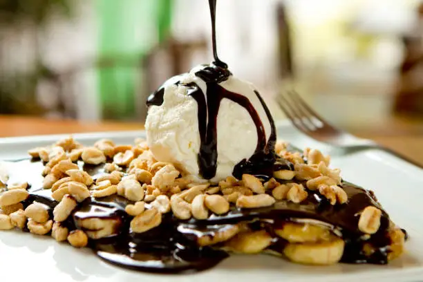 Chocolate crepe and vanilla ice cream, banana slices and peanuts toppings. Syrup dripping from top. Tasty, sweet gourmet dessert served on wood table, closeup view, blurry background. French creperie.