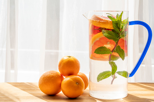 A blue handle glass carafe (pitcher) in sunlight with high PH level detox water to healthy life and boost the immune system to fight any illness, plus corona and other viruses. Ingredients: grapefruit, strawberry, orange and mint for a best aroma