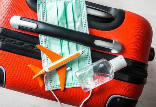 Photo of Flat lay view of medical face mask with airplane figurine on traveling baggage suitcase with small portable hand sanitizer bottle. Traveling during coronavirus outbreak concept. Year 2020.