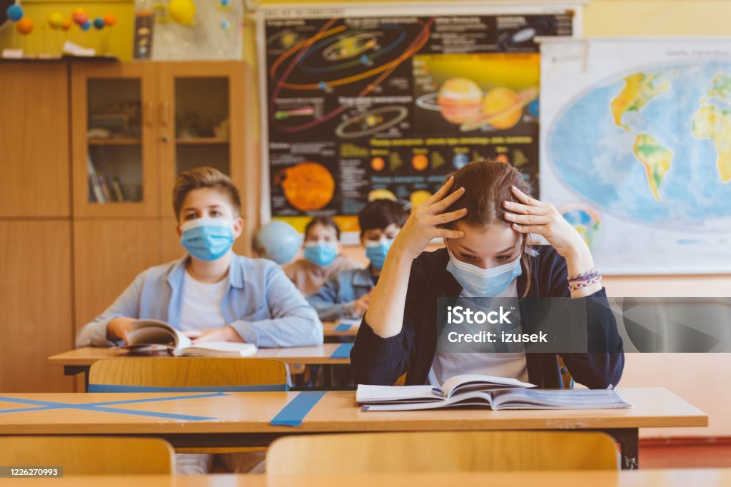 Worried high school girl student at school wearing N95 Face masks High school students at school, wearing N95 Face masks. Worried teenage girl sitting at the school desk, holding head in hands. Education Stock Photo