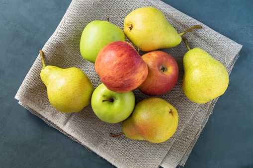 A few fresh bright apples and pears lie on a simple coarse rustic napkin on a dark blue background. Healthy eating. Horizontal orientation, view from above, selective focus.