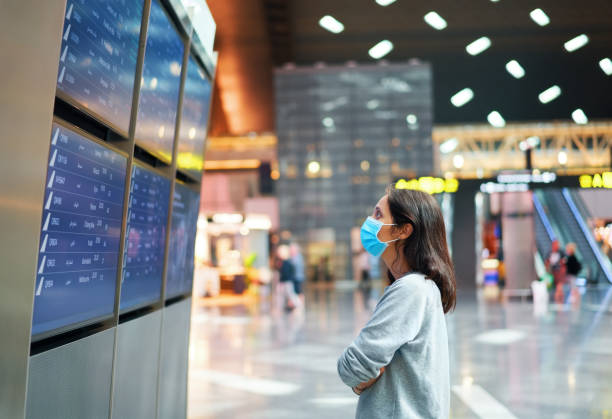 Woman in virus protection face mask looking at information board checking her flight in international airport Woman in virus protection face mask looking at information board checking her flight in international airport. Departure board, flight status cancellation photos stock pictures, royalty-free photos & images