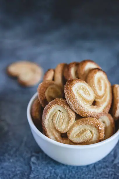 Photo of Palmier Puff Pastry. Delicious french palmier cookies with sugar in a white bowl on dark background.