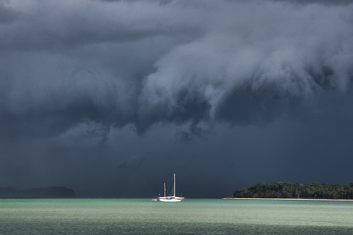 Extreme weather dark Virga thunder clouds with torrential rain are forming above the sea.  Below is a small, white sailboat.  These weather events are also known as tornado, cyclone or hurricane, are unfortunately becoming more common around the world due to Global Warming and Climate Change.  These weather patterns are violent and dangerous, often claiming numerous lives in the areas which they occur.  Location is Andaman sea, Thailand.