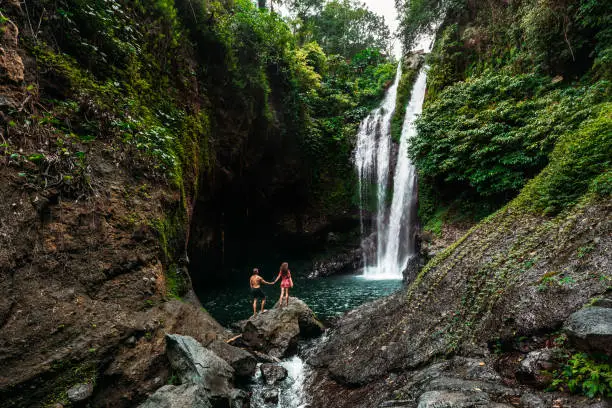 Photo of Lovers at the waterfall, rear view. Couple admiring a beautiful waterfall in Indonesia. Couple on vacation in Bali. Honeymoon trip. The couple is traveling in Asia. Vacation on the island of Bali