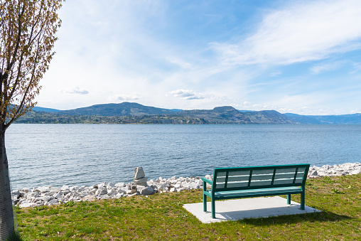 Scenic view of empty park bench, Okanagan Lake, mountains, and blue sky on sunny afternoon at Naramata Wharf Park in Naramata, British Columbia, Canada in the Okanagan Valley