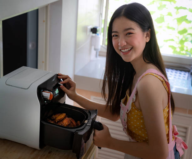 自宅で彼女の台所でエアフライヤーマシンでフライドチキンを調理アジアの女の子 - w2 ストックフォトと画像
