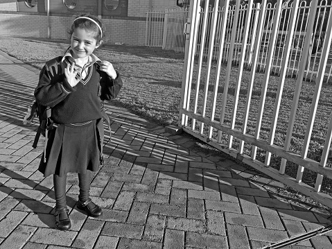 Young school girl (age 6) wearing school uniform and carrying a bag waving goodbye before entering school.
