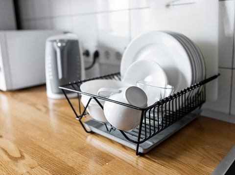 Budget and lightweight antimicrobial dish drainer with drain board at modern scandinavian kitchen. Dish rack holds many dishes and cups against wooden countertop, white wall tiles, sink and faucet
