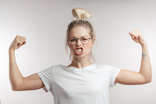 I can everything. Happy young blonde woman in white t-shirt showing biceps on her arms. Emotional portrait with white background of expressive girl with blonde hair bun and spectacles.