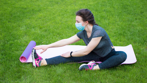 居心地の良い-19の間にスポーツ。顔の医療マスクを身に着けている若い女性は、屋外でトレーニングフィットネスを練習する足を伸ばします。ストレッチ運動をしている草の上に座って美し� - crossfit exercising sports training outdoors ストックフォトと画像