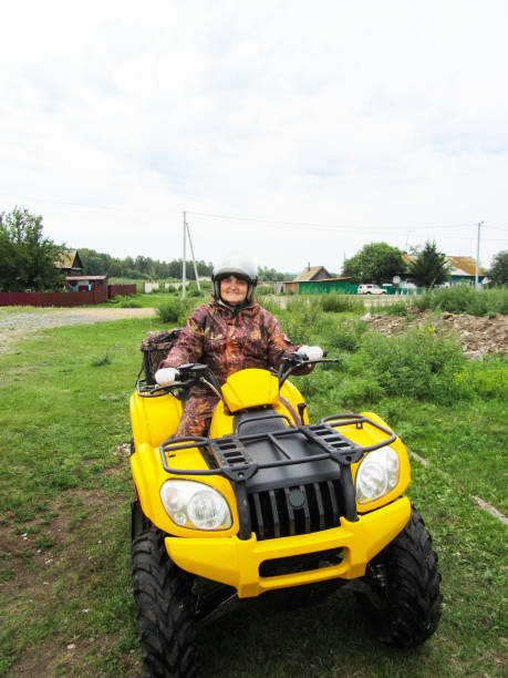 eine zufriedene dicke ältere frau im helm reitet mit einem gelben atv durch das dorf. sport, erholung, unterhaltung - off road vehicle quadbike quadruplet motocross stock-fotos und bilder