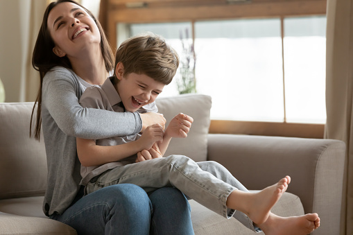 Excited young Caucasian mom sit on couch in living room play with happy little preschooler son, smiling loving mother or nanny have fun engaged in funny activity with overjoyed small boy child