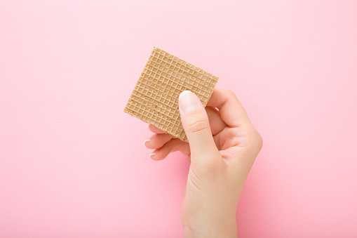 Young woman hand holding brown wafer on light pink table background. Crispy sweet snack. Closeup. Top down view.