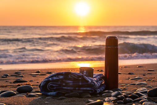 Thermos of hot tea and a soft plaid on a sandy beach early in the morning at sunrise
