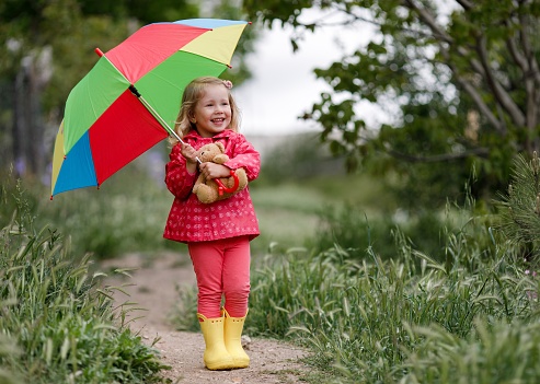 Happy funny beautiful blonde with blue eyes child with rainbow colorful umbrella in the autumn park. Girl kid playing on the nature outdoors. Family walk in the may forest green trees leaves.Girl hiding under a umbrella from spring rain.