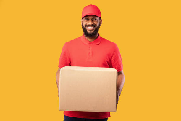 istock Smiling Courier Offering Cardboard Box To Camera Over Yellow Background 1226380417