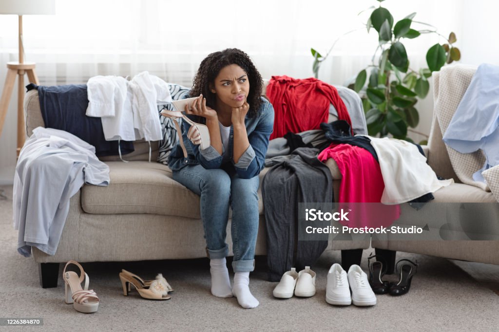 Unsuccessful Online Shopping. Disappointed Woman Sitting Beside Piles Of Unfitting Clothes Unsuccessful Online Shopping. Disappointed Pensive Black Woman Sitting On Couch Beside Piles Of Unfitting Clothes And Shoes Ordered In Internet Messy Stock Photo