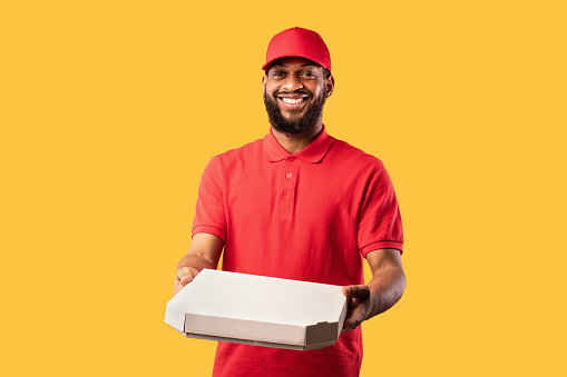 African Courier Guy Delivering Pizza Box Standing In Studio On Yellow Background. Pizzeria Delivery Service.