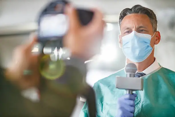 Photo of Male doctor giving an interview in front of the camera in the hospital.