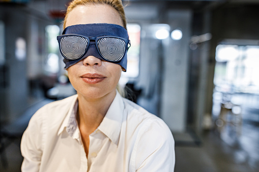 Blindfolded female entrepreneur with hypnotic eyeglasses unable to see the reality in the office.