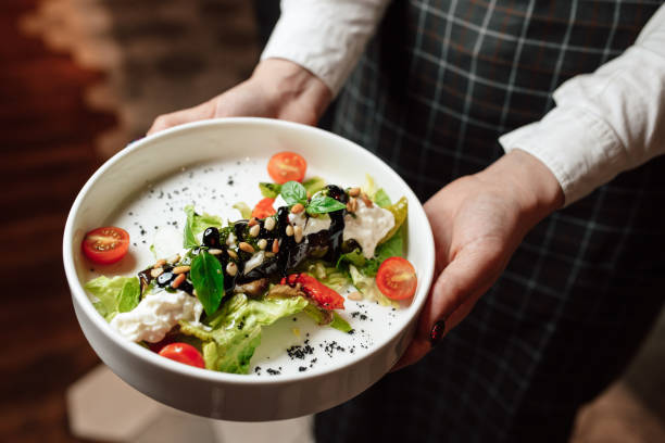 il cameriere tiene in mano un piatto bianco con un piatto, primo piano delle mani. insalata con pomodorini, basilico e pinoli - piatto di portata foto e immagini stock