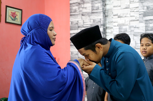 A Muslim young man is giving 'salam' (Muslim way of greeting or asking for forgiveness) to his mother at home in Malaysia. She is a Thai ethnicity and convert to Islam before marriage, wearing 'telekung' (female praying robe) after evening prayers.