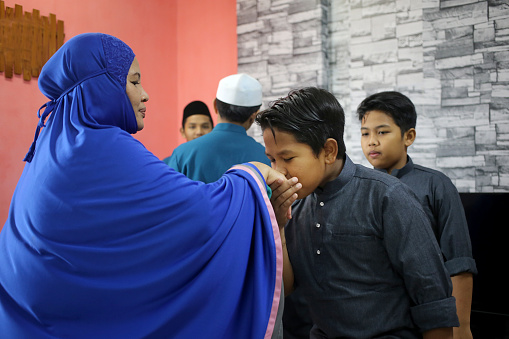 A Muslim young man is giving 'salam' (Muslim way of greeting or asking for forgiveness) to his mother at home in Malaysia. She is a Thai ethnicity and convert to Islam before marriage, wearing 'telekung' (female praying robe) after evening prayers.