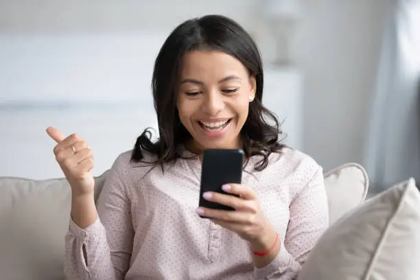 Photo of African female holds smartphone feels overjoyed showing thumbs up